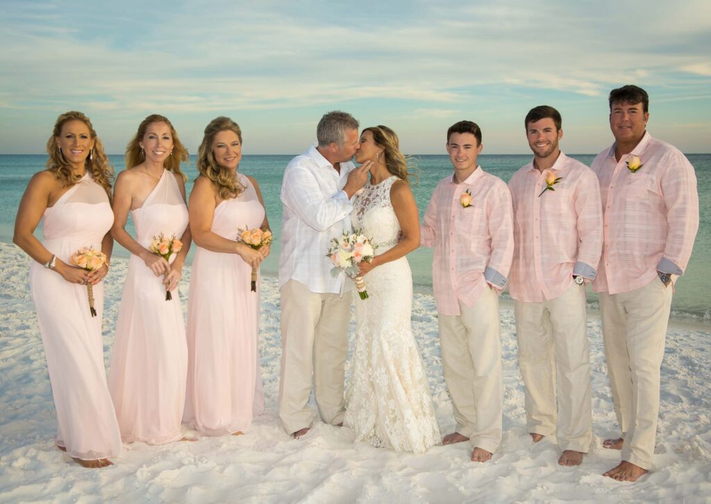 Groom kissing bride on the beach with bridesmaids and groomsmen on either side. A sunset beach wedding photo.